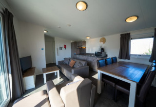 Indvendigt billede af en hyggelig lodge med sofa, spisebord, tekøkken og vinduer med gardiner.