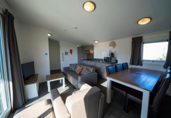 Interieur van een gezellige lodge met bank, eettafel, kitchenette en ramen met gordijnen.