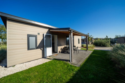 Modernes Lodge mit überdachter Terrasse und Gartenmöbeln, umgeben von grünem Rasen unter blauem Himmel.