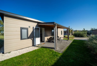 Modernes Lodge mit überdachter Terrasse und Gartenmöbeln, umgeben von grünem Rasen unter blauem Himmel.