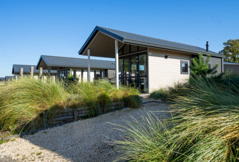 Foto einer modernen Lodge mit großen Fenstern, Kiesweg und hohem Gras unter klarem, blauem Himmel.