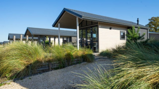 Foto van een moderne lodge met grote ramen, een grindpad en hoge siergrassen onder een blauwe hemel.