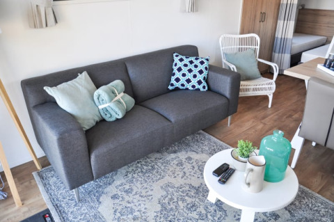Sala de lodge moderna con sofá gris, cojines, mesa blanca, plantas, tazas y decoración elegante.