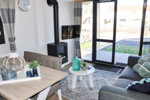 Lichte en moderne lodge woonkamer met bank, eettafel, haard, tv en groot raam met tuinzicht.