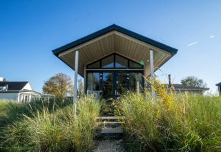 Photo d'un pavillon moderne aux grandes baies vitrées, entouré de hautes herbes sous un ciel bleu clair.