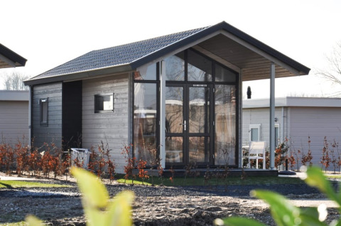 Modern wooden lodge with large glass windows and a small porch, surrounded by shrubs and nearby lodges.