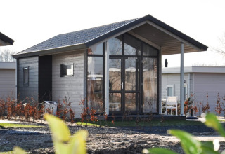 Modern wooden lodge with large glass windows and a small porch, surrounded by shrubs and nearby lodges.