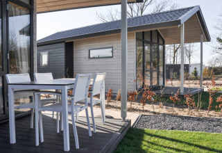 Udendørs terrasse ved en moderne lodge med hvide havemøbler og udsigt til en anden lodge i baggrunden.