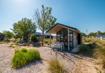 Moderne lodge med terrasse og udendørs siddeplads, omgivet af grøn beplantning og blå himmel.