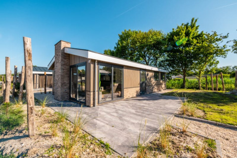 Lodge moderne avec murs en verre et terrasse, entouré de nature et d’arbres sous un ciel bleu clair.