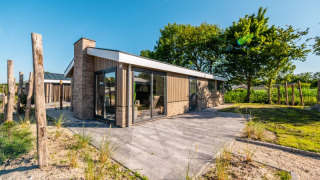 Cabaña moderna con paredes de cristal y terraza, rodeada de naturaleza y árboles bajo cielo despejado.