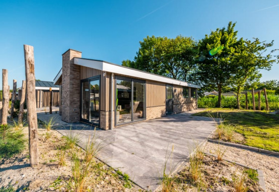 Lodge moderne avec murs en verre et terrasse, entouré de nature et d’arbres sous un ciel bleu clair.