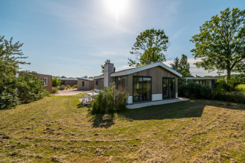 Lodge moderne avec grandes baies vitrées, pelouse verte et arbres sous un ciel bleu ensoleillé.