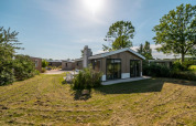 Moderno lodge con grandes ventanales, césped verde y árboles bajo un cielo azul y soleado.