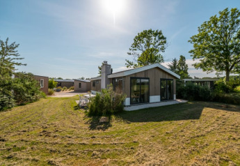 Lodge moderne avec grandes baies vitrées, pelouse verte et arbres sous un ciel bleu ensoleillé.