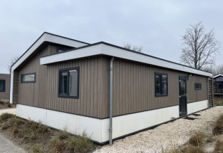 Un pavillon en bois moderne avec des cadres de fenêtres noirs, installé sur un chemin de gravier entouré d’herbe.