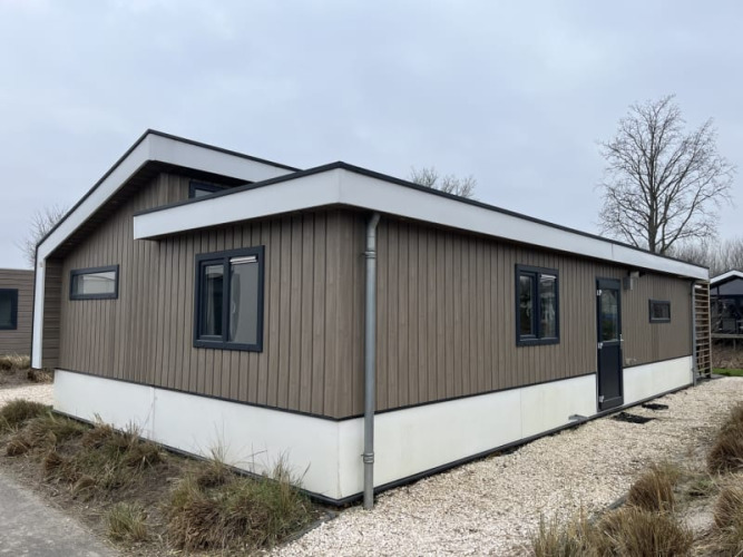 Una moderna cabaña de madera con marcos de ventana negros ubicada en un sendero de grava entre césped y árboles.