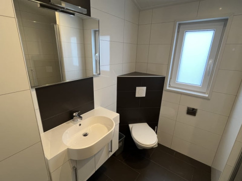 Baño moderno en una cabaña, con inodoro de pared, lavabo blanco, espejo grande y ventana esmerilada.
