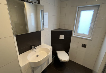 Bagno moderno in un lodge con wc sospeso, lavabo bianco, grande specchio e finestra con vetro satinato.