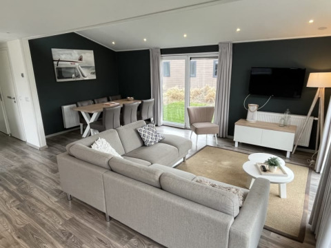 Salon de lodge moderne avec canapé d’angle gris, grande baie vitrée, table à manger et télévision murale.