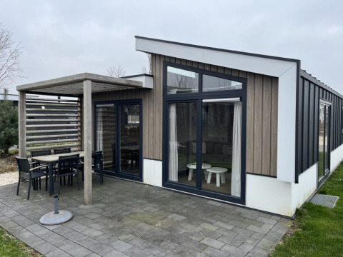 Lodge moderne avec grandes baies vitrées, coin repas extérieur couvert et terrasse pavée entourée de pelouse.