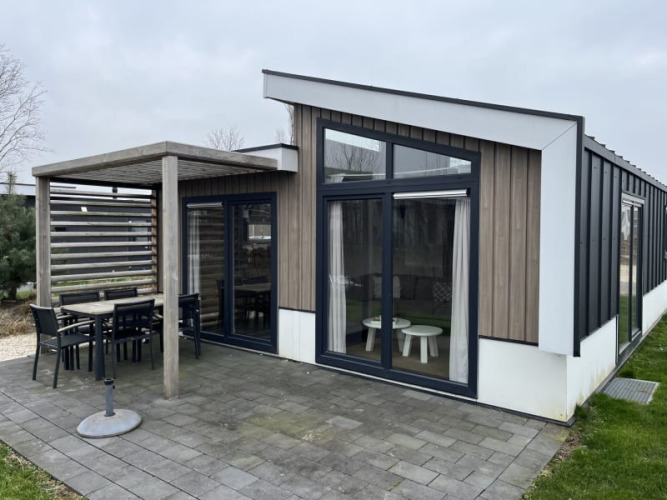 Moderna cabaña con grandes ventanales, comedor exterior cubierto y patio rodeado de césped verde.