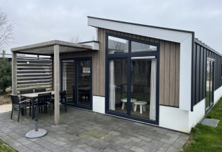 Moderna cabaña con grandes ventanales, comedor exterior cubierto y patio rodeado de césped verde.