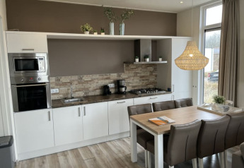 Cocina moderna en una cabaña, mesa de comedor, electrodomésticos y grandes ventanales soleados.