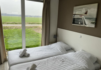 Chambre lumineuse d'un lodge avec deux lits simples, grandes fenêtres et vue sur un champ vert.