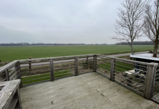 Vue depuis une terrasse en bois d’un lodge donnant sur des champs ouverts et des arbres sans feuilles sous ciel gris.