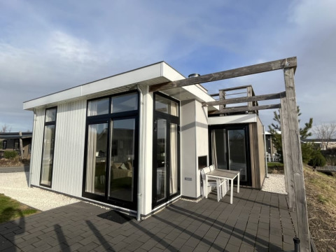 Lodge moderne avec grandes baies vitrées, terrasse et pergola en bois dans un cadre naturel sous ciel bleu.