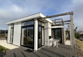 Lodge moderne avec grandes baies vitrées, terrasse et pergola en bois dans un cadre naturel sous ciel bleu.