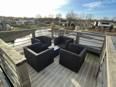 Terrasse sur le toit d’un lodge avec quatre fauteuils en osier noir, table et vue sur chalets et canal.