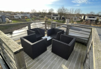 Terrasse sur le toit d’un lodge avec quatre fauteuils en osier noir, table et vue sur chalets et canal.
