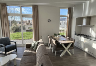 Salon moderne et lumineux d'un lodge avec grandes fenêtres, vue sur jardin, table à manger et cuisine ouverte.