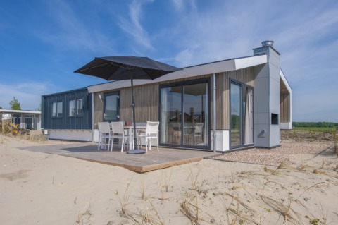 Moderne Lodge mit großen Fenstern, Terrasse und Sonnenschirm auf sandigem Gelände unter blauem Himmel.