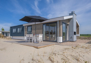 Moderne Lodge mit großen Fenstern, Terrasse und Sonnenschirm auf sandigem Gelände unter blauem Himmel.