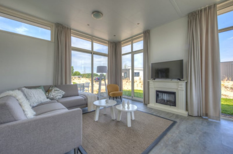 Salon moderne de lodge avec grandes baies vitrées, canapé gris, fauteuil jaune, cheminée, TV et vue jardin.