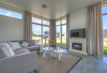 Salon moderne de lodge avec grandes baies vitrées, canapé gris, fauteuil jaune, cheminée, TV et vue jardin.