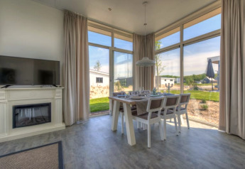 Bright lodge interior with large windows, dining table, TV on a fireplace, and scenic outdoor landscape views.