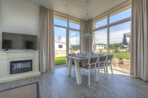 Interior luminoso de una cabaña con grandes ventanales, mesa de comedor, televisor y vistas al paisaje exterior.