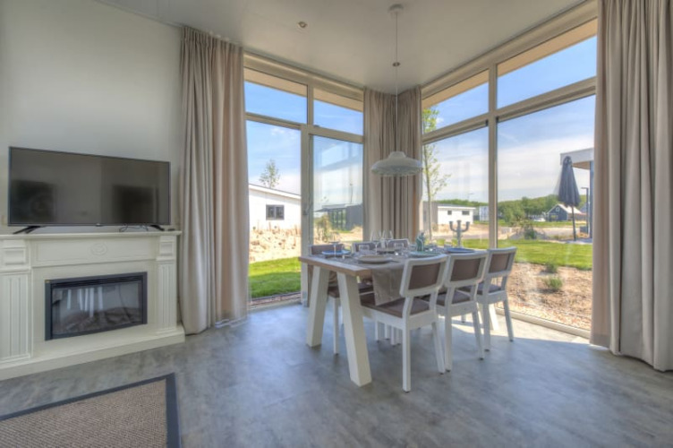 Interior luminoso de una cabaña con grandes ventanales, mesa de comedor, televisor y vistas al paisaje exterior.