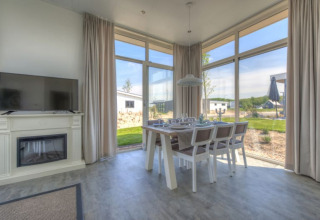 Intérieur lumineux d'un lodge avec grandes fenêtres, table à manger, télévision et vue sur le paysage extérieur.