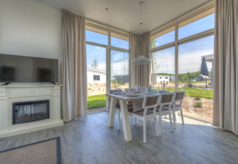 Interior luminoso de una cabaña con grandes ventanales, mesa de comedor, televisor y vistas al paisaje exterior.