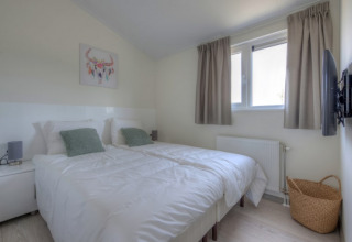 Chambre moderne et lumineuse d’un lodge avec deux lits simples, coussins verts, tableau mural et TV.