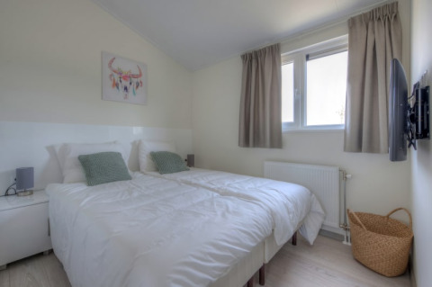 Chambre moderne et lumineuse d’un lodge avec deux lits simples, coussins verts, tableau mural et TV.