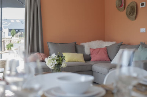 Salon de lodge avec mur orange, canapé d’angle gris, coussins colorés et vue sur la terrasse extérieure.