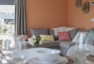 Sala de estar de lodge con pared naranja, sofá gris en forma de L, cojines de colores y vista a la terraza.