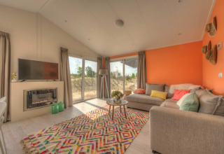 Salon de lodge moderne avec mur orange, grandes fenêtres, canapé beige, TV et tapis coloré au sol.