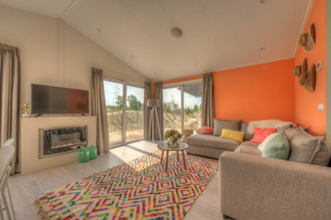 Salon de lodge moderne avec mur orange, grandes fenêtres, canapé beige, TV et tapis coloré au sol.